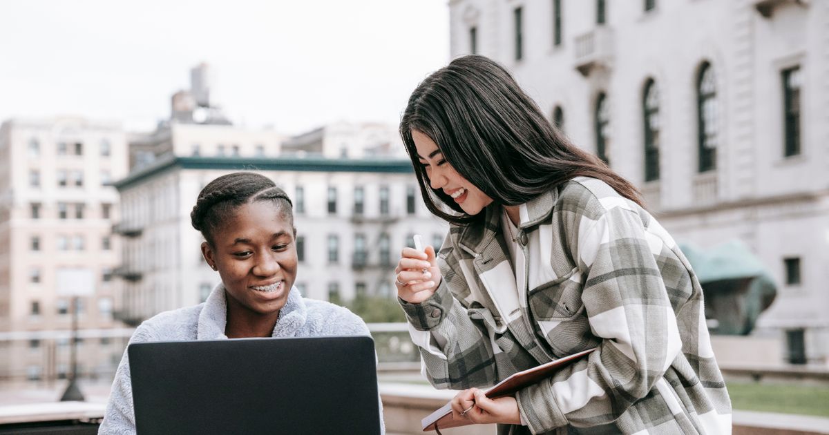 Twee jonge vrouwen kijken samen glimlachend naar een laptop buiten bij een universiteitsgebouw